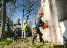 Construction workers dressed in safety gear demolishing a brick wall with heavy machinery outdoors.